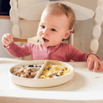 One-year-old baby sitting in a high chair, feeding herself yogurt with a spoon, with a divided plate of scrambled eggs, toast pieces, and blueberries — realistic toddler breakfast idea.