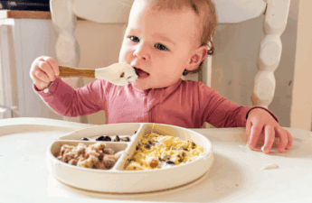 One-year-old baby sitting in a high chair, feeding herself yogurt with a spoon, with a divided plate of scrambled eggs, toast pieces, and blueberries — realistic toddler breakfast idea.