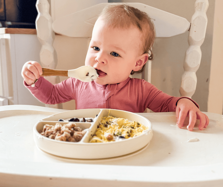 One-year-old baby sitting in a high chair, feeding herself yogurt with a spoon, with a divided plate of scrambled eggs, toast pieces, and blueberries — realistic toddler breakfast idea.