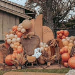 Outdoor fall baby shower setup featuring a terracotta, beige, and burnt orange balloon garland surrounding a rattan peacock chair. Pampas grass and dried palm fronds are arranged throughout, with curved neutral backdrops behind the chair and a cozy baby blanket draped over the seat. The decor creates a warm, boho autumn vibe.