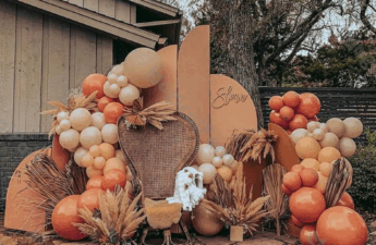 Outdoor fall baby shower setup featuring a terracotta, beige, and burnt orange balloon garland surrounding a rattan peacock chair. Pampas grass and dried palm fronds are arranged throughout, with curved neutral backdrops behind the chair and a cozy baby blanket draped over the seat. The decor creates a warm, boho autumn vibe.