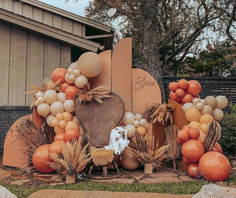 Outdoor fall baby shower setup featuring a terracotta, beige, and burnt orange balloon garland surrounding a rattan peacock chair. Pampas grass and dried palm fronds are arranged throughout, with curved neutral backdrops behind the chair and a cozy baby blanket draped over the seat. The decor creates a warm, boho autumn vibe.