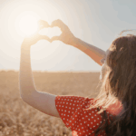 Woman in red dress holding her hands in the shape of a heart toward the setting sun in a golden field