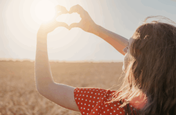 Woman in red dress holding her hands in the shape of a heart toward the setting sun in a golden field