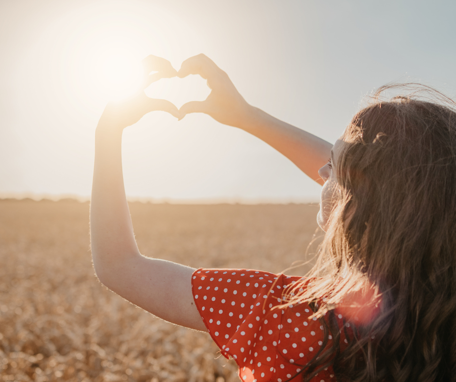 Woman in red dress holding her hands in the shape of a heart toward the setting sun in a golden field