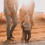 Toddler boy walking outdoors in warm golden sunlight, holding a jacket, with two adults walking behind him in a field during autumn.