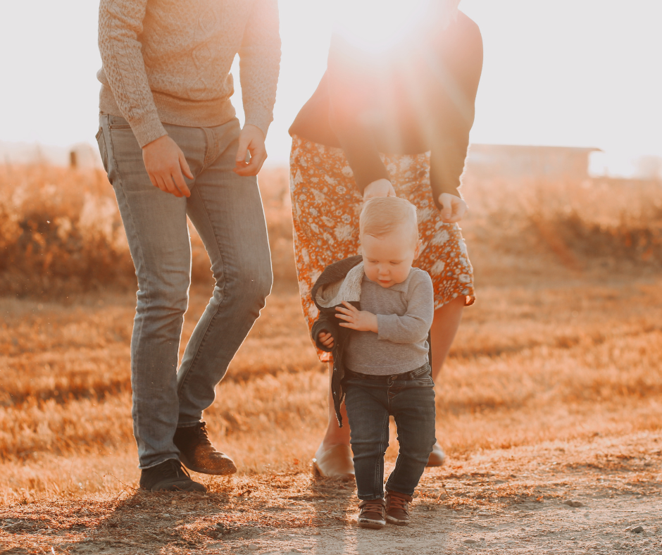 Toddler boy walking outdoors in warm golden sunlight, holding a jacket, with two adults walking behind him in a field during autumn.