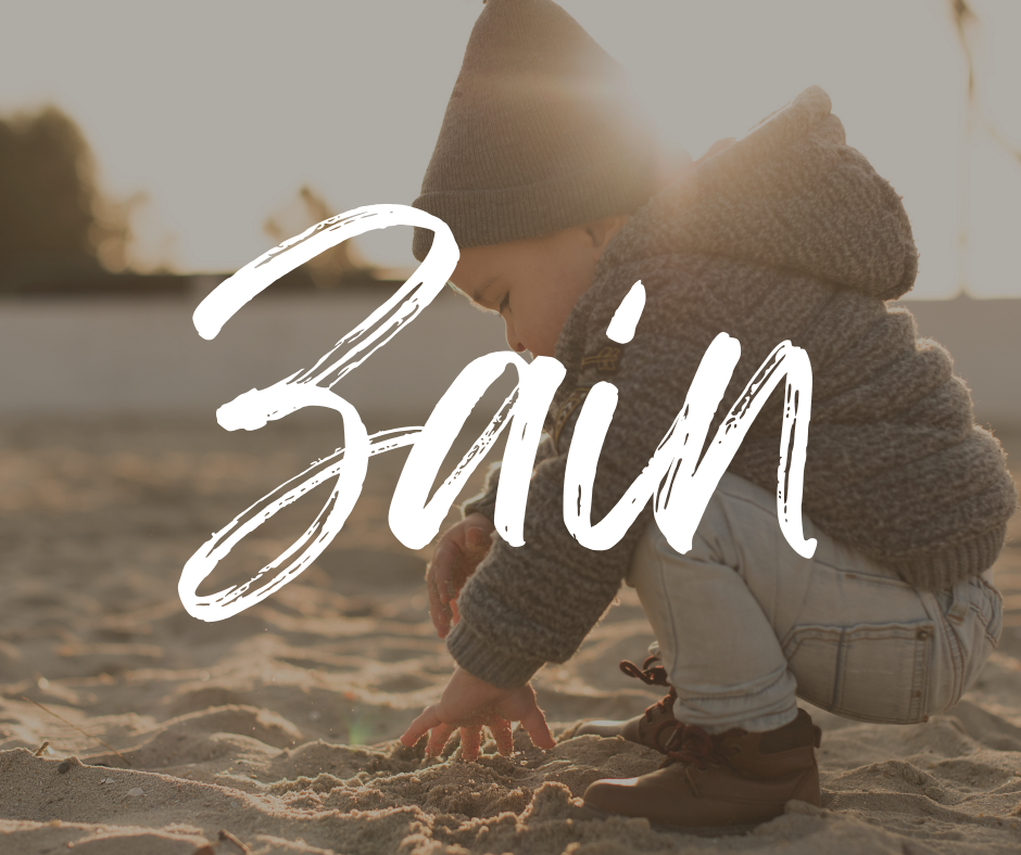 Toddler wearing a knit beanie, gray sweater, and light jeans squatting to play in the sand at sunset, with the name “Zain” written in large white script across the image.