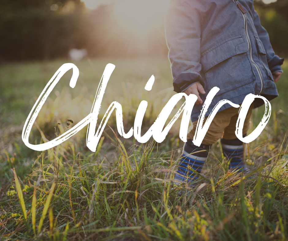 Close-up of a toddler standing in a grassy field wearing a blue jacket, tan pants, and striped rain boots, with the name “Chiaro” written in large white script across the image.