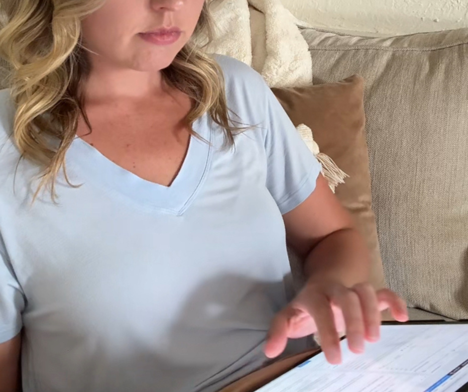 Woman sitting on a beige couch using a tablet to work on her mom blog, wearing a light blue V-neck shirt.