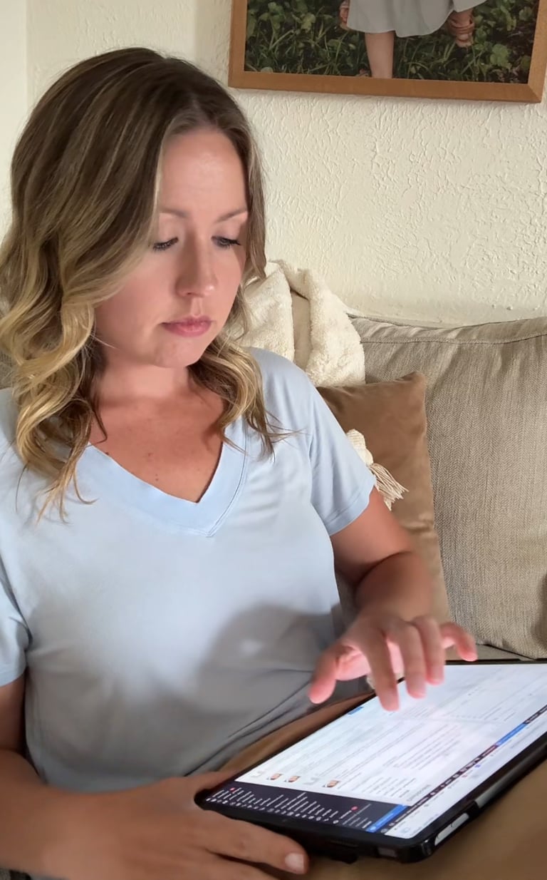 Woman sitting on a beige couch using an iPad, focused on the screen while scrolling or tapping with her finger. A framed photo is visible on the wall behind her.