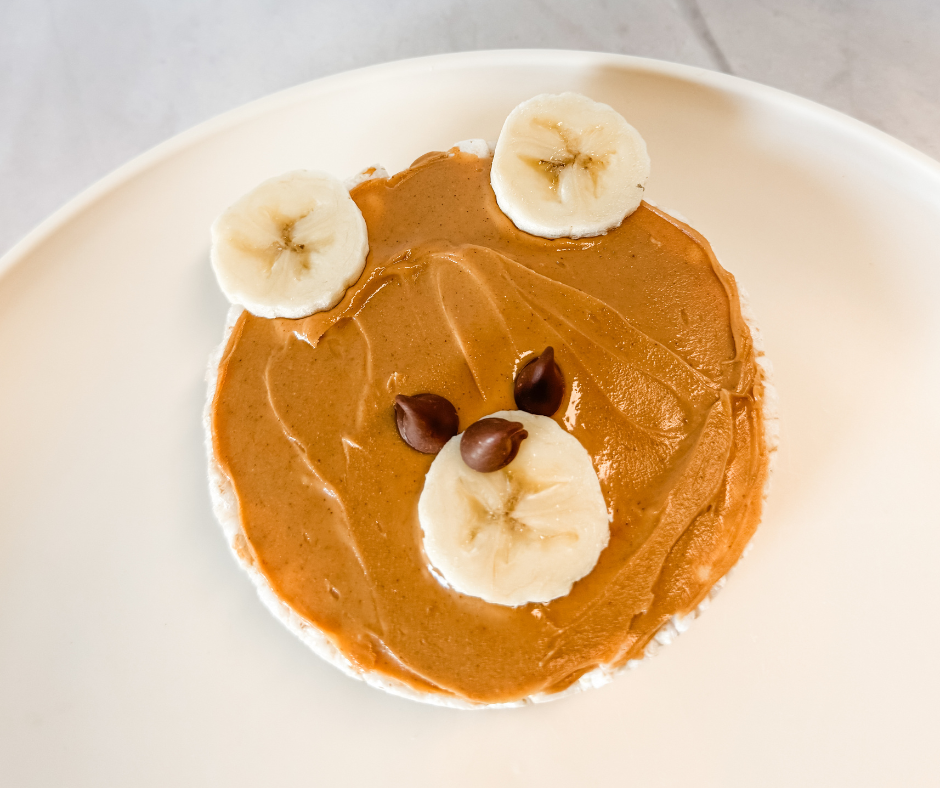 Peanut butter bear snack made with a rice cake, banana slices for ears and snout, and chocolate chips for the face. A fun and healthy snack idea for kids.