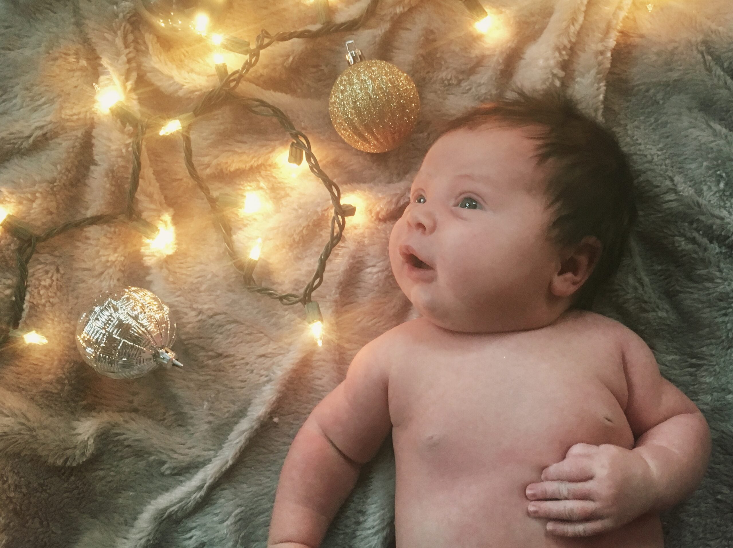 Newborn baby lying on a fuzzy gray blanket with glowing Christmas lights and sparkly ornaments, looking up in wonder.