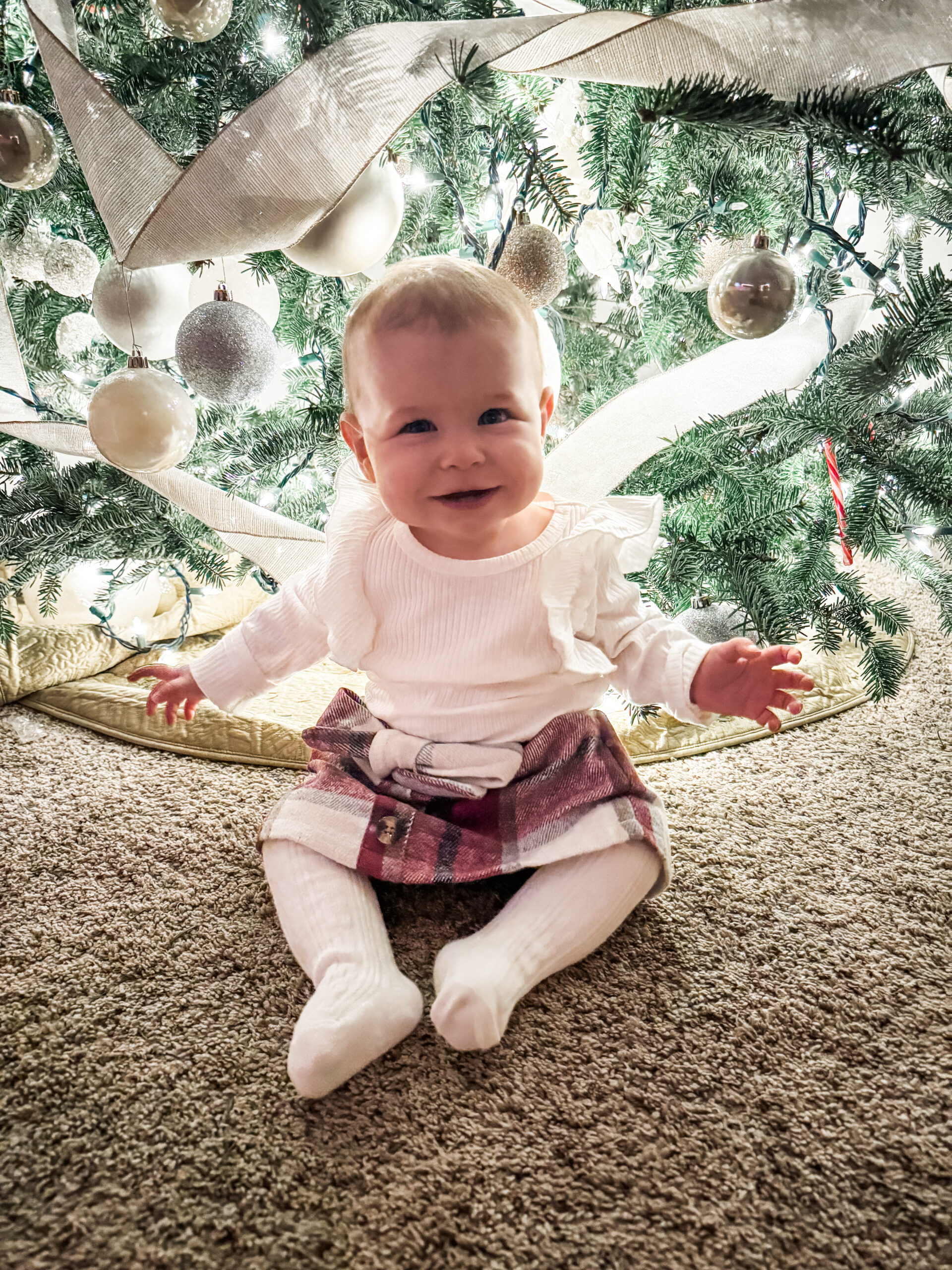 Smiling baby sitting in front of a decorated Christmas tree with white ornaments and ribbon, wearing a plaid skirt and white top.