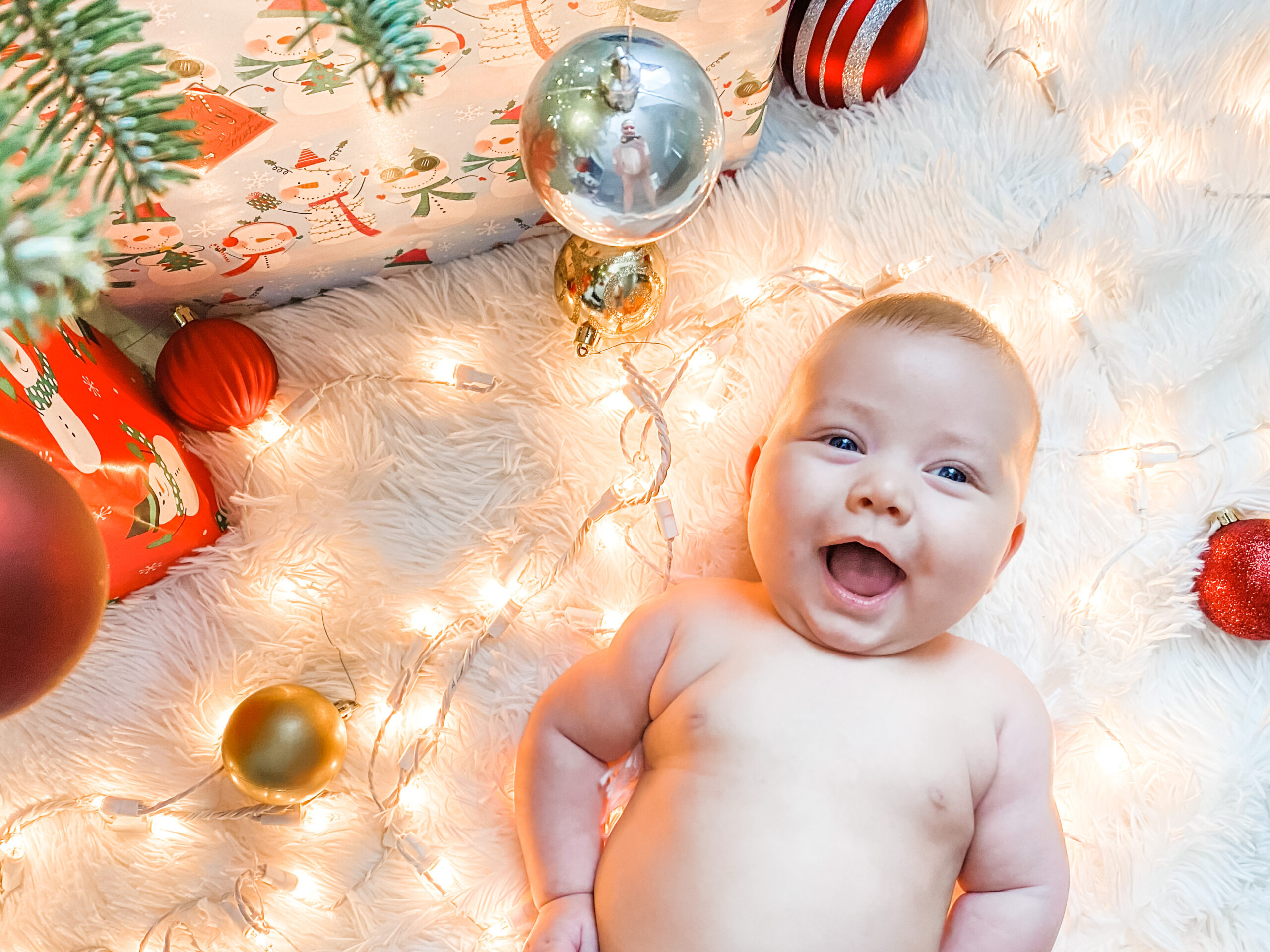 Baby lying on a soft white blanket surrounded by Christmas lights, ornaments, and wrapped presents with a Christmas tree branch in the corner.