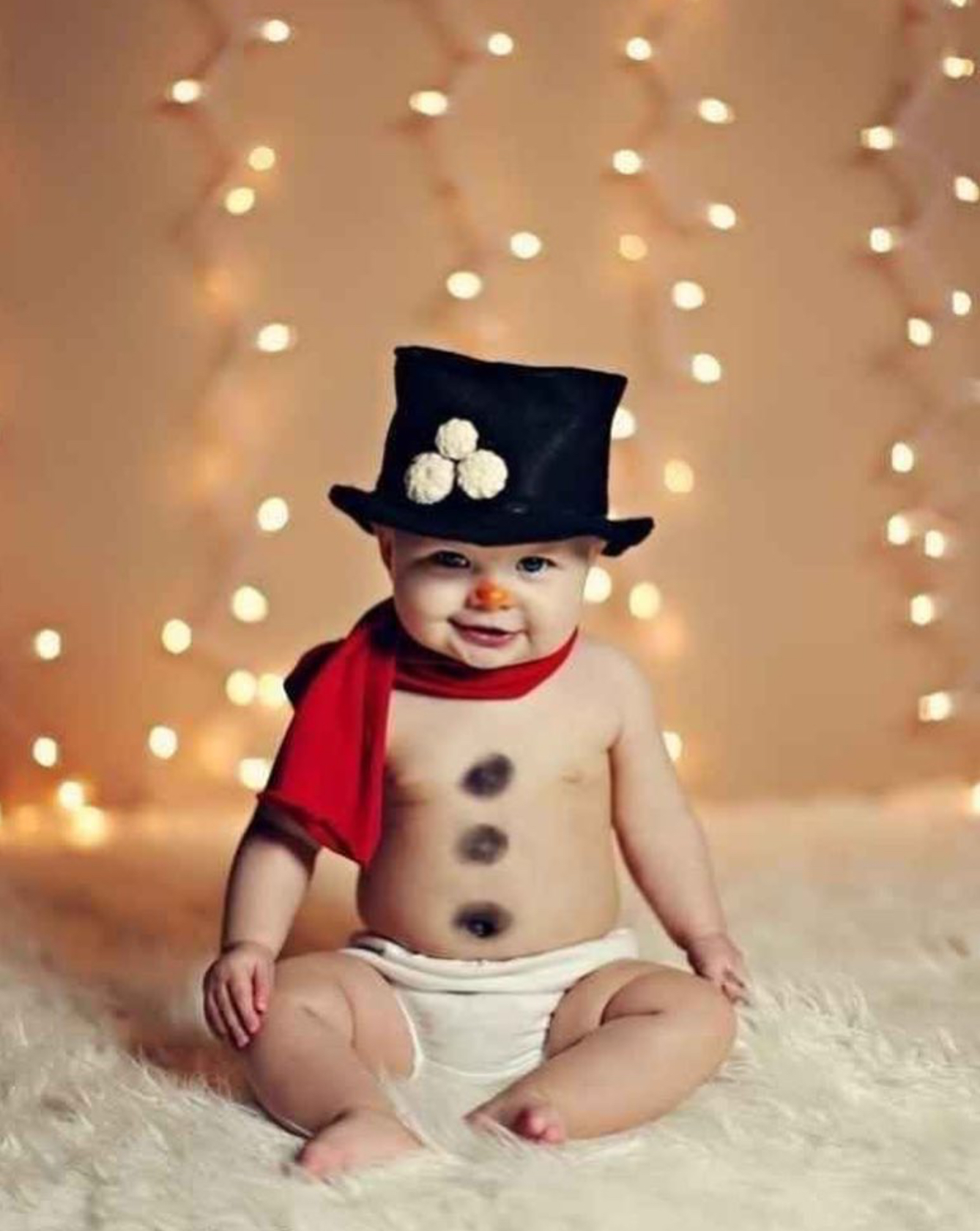 Baby on white blanket with Christmas lights behind him. He’s dressed in a black top hat and red scarf with 3 black circles on his belly to look like a snowman.
