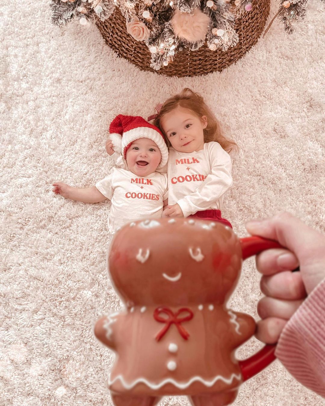 Two siblings lying on a soft white rug beneath a decorated Christmas tree, wearing matching “Milk + Cookies” shirts; the baby wears a Santa hat while a hand in the foreground holds a cute gingerbread man mug.