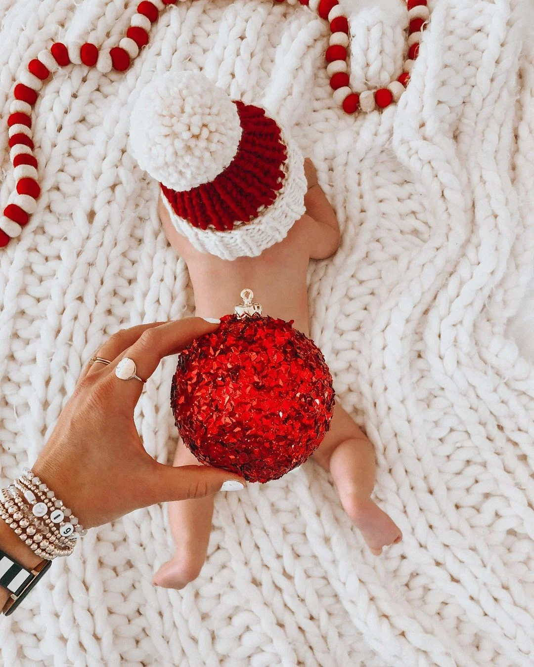 Newborn baby lying on a chunky white knit blanket wearing a red and white knit hat, with a parent holding a large red Christmas ornament in the foreground; red and white garland decorates the blanket.