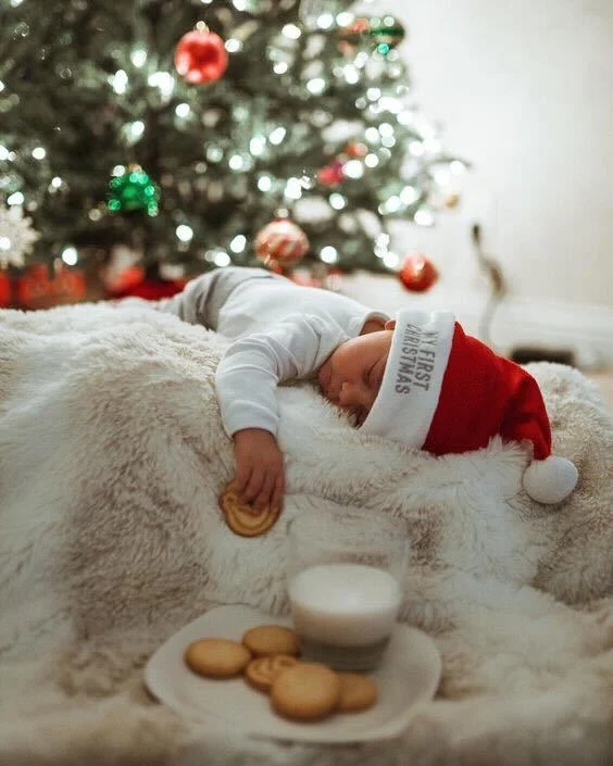 Baby wearing a “My First Christmas” Santa hat asleep on a fluffy blanket in front of a lit Christmas tree, holding a cookie, with a plate of cookies and milk in the foreground.