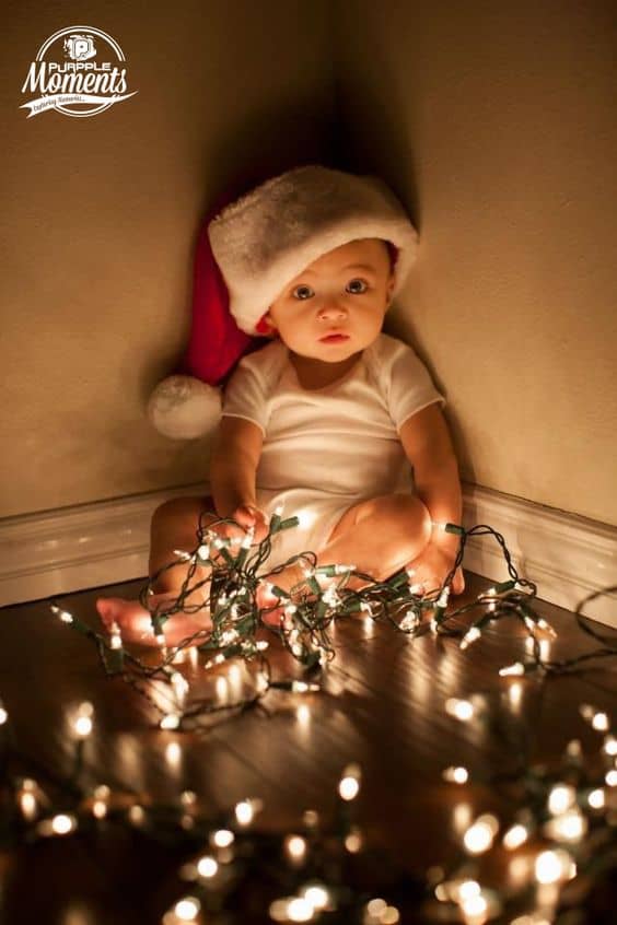 Baby wearing a Santa hat and white onesie sitting in a corner on hardwood floor, surrounded by glowing Christmas lights, looking up with wide eyes.