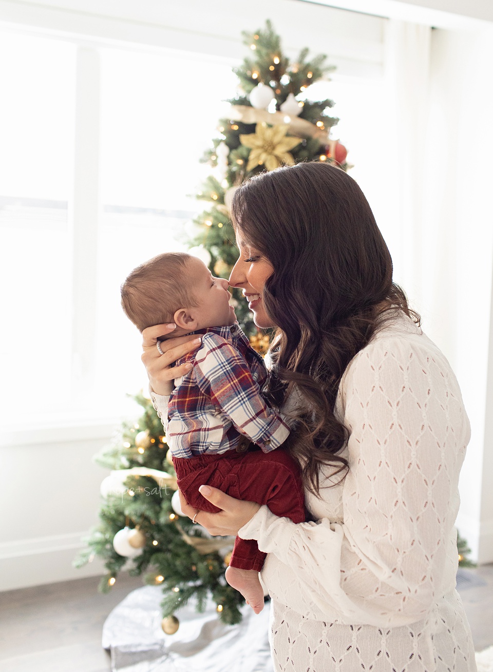 Mom holding her baby in front of a decorated Christmas tree, touching noses and smiling in soft natural light.