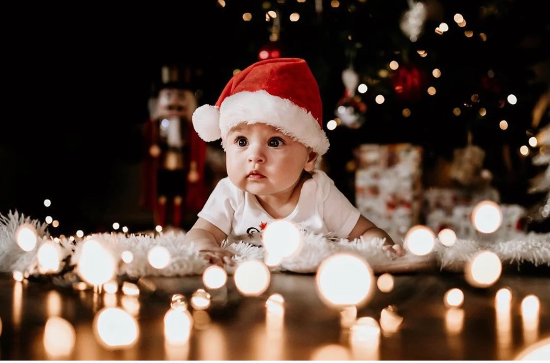 Baby wearing a Santa hat lying on a fluffy rug with glowing Christmas lights in the foreground and a decorated tree and wrapped gifts behind.