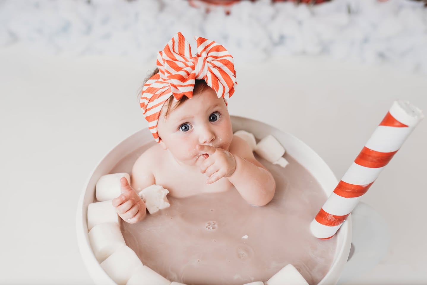 Baby sitting in a round tub of warm cocoa-colored water with large marshmallows, wearing a red-and-white striped headband, with a giant striped straw prop.