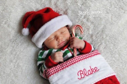 Newborn baby wearing a Santa hat and striped Christmas pajamas, tucked inside a personalized stocking and holding a candy cane while sleeping on a soft white blanket.
