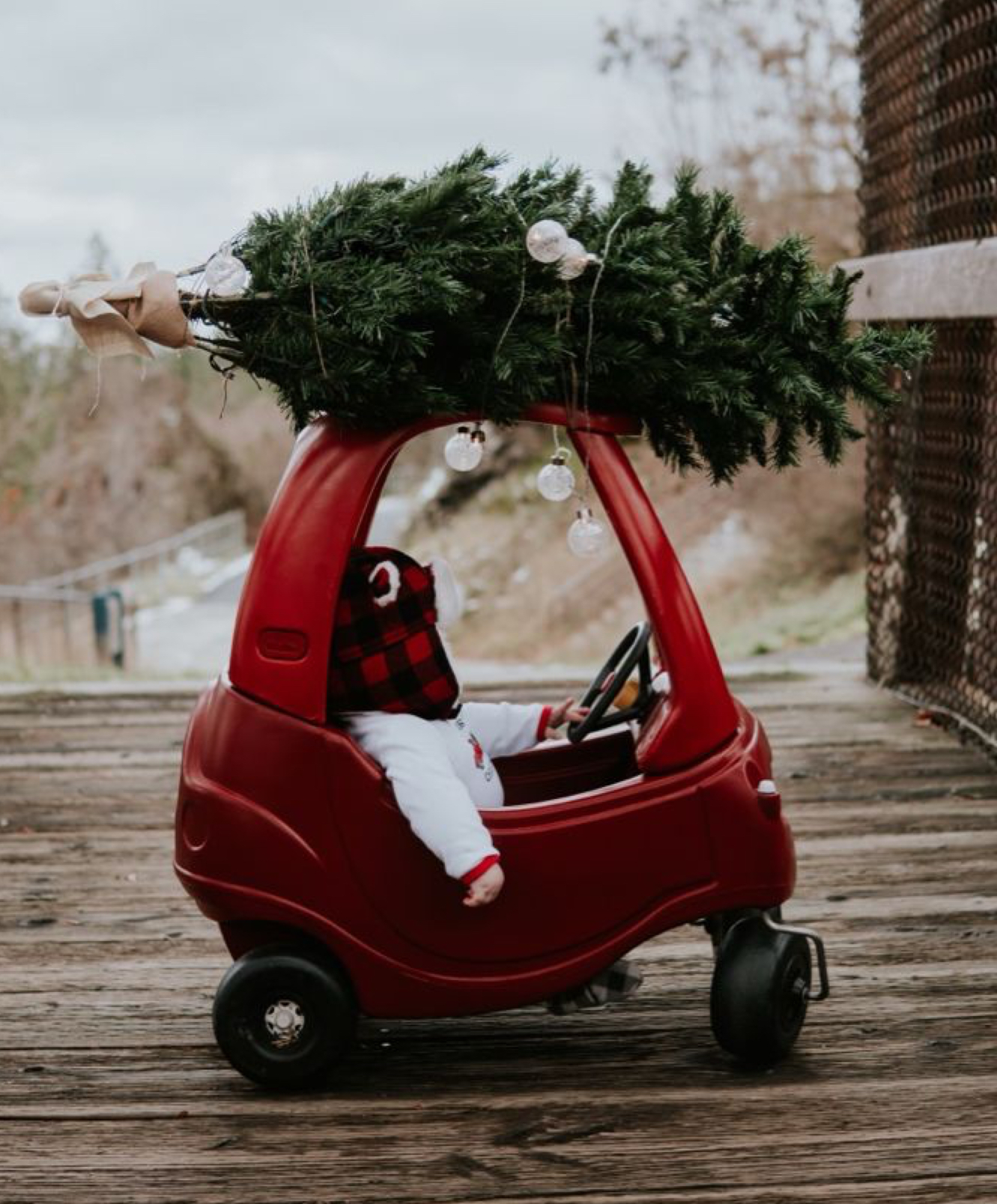 Baby in a red toy car wearing a buffalo plaid winter hat, with a small Christmas tree tied to the roof, sitting on a wooden bridge outdoors.