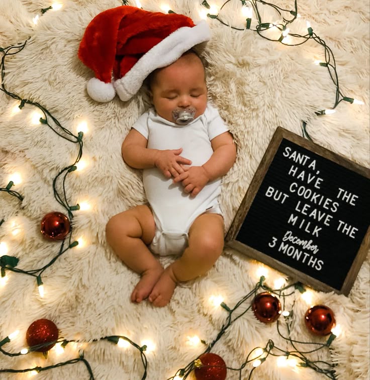 Sleeping baby wearing a Santa hat on a fluffy cream blanket, surrounded by Christmas lights and red ornaments, next to a letter board that says “Santa, have the cookies but leave the milk — December, 3 months.”