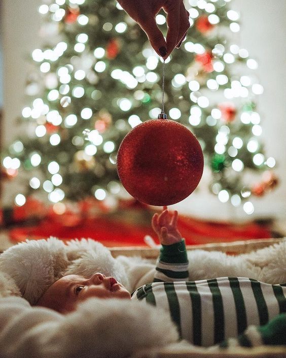 Baby lying on a cozy blanket in front of a lit Christmas tree, reaching up toward a large red ornament being held above them.