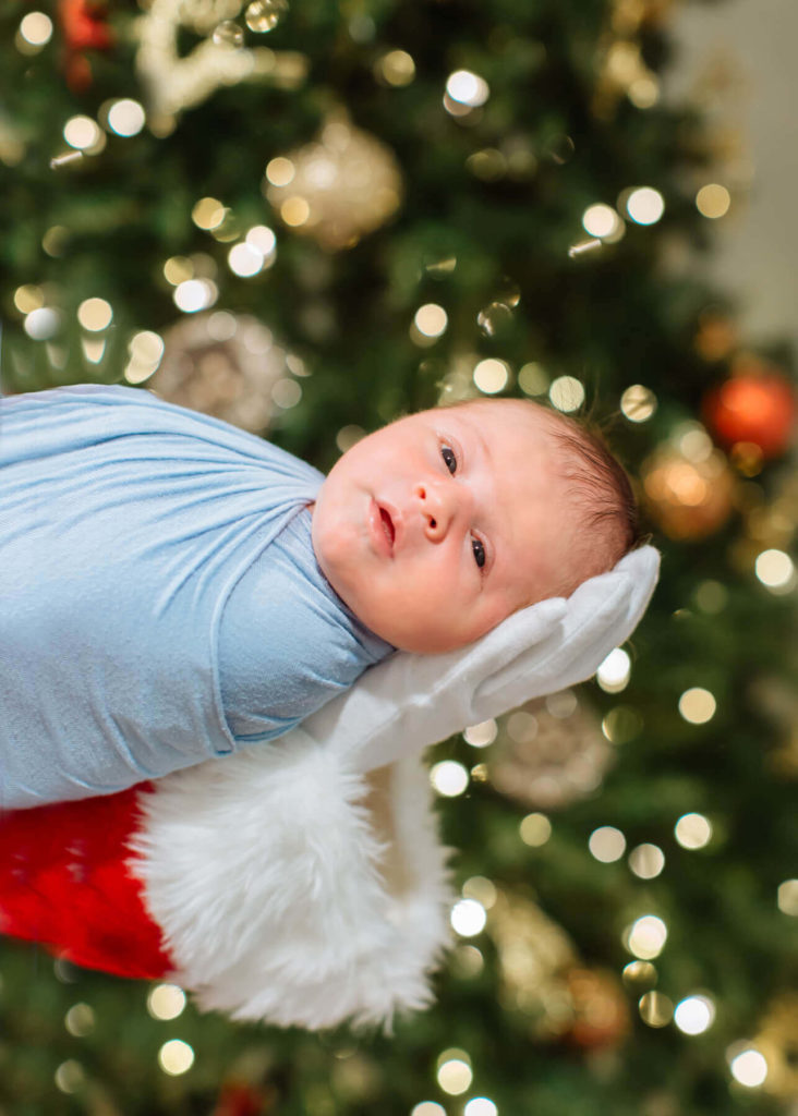 Newborn baby wrapped in a blue swaddle being held horizontally by someone wearing a Santa suit and white glove, with a decorated Christmas tree glowing in the background.