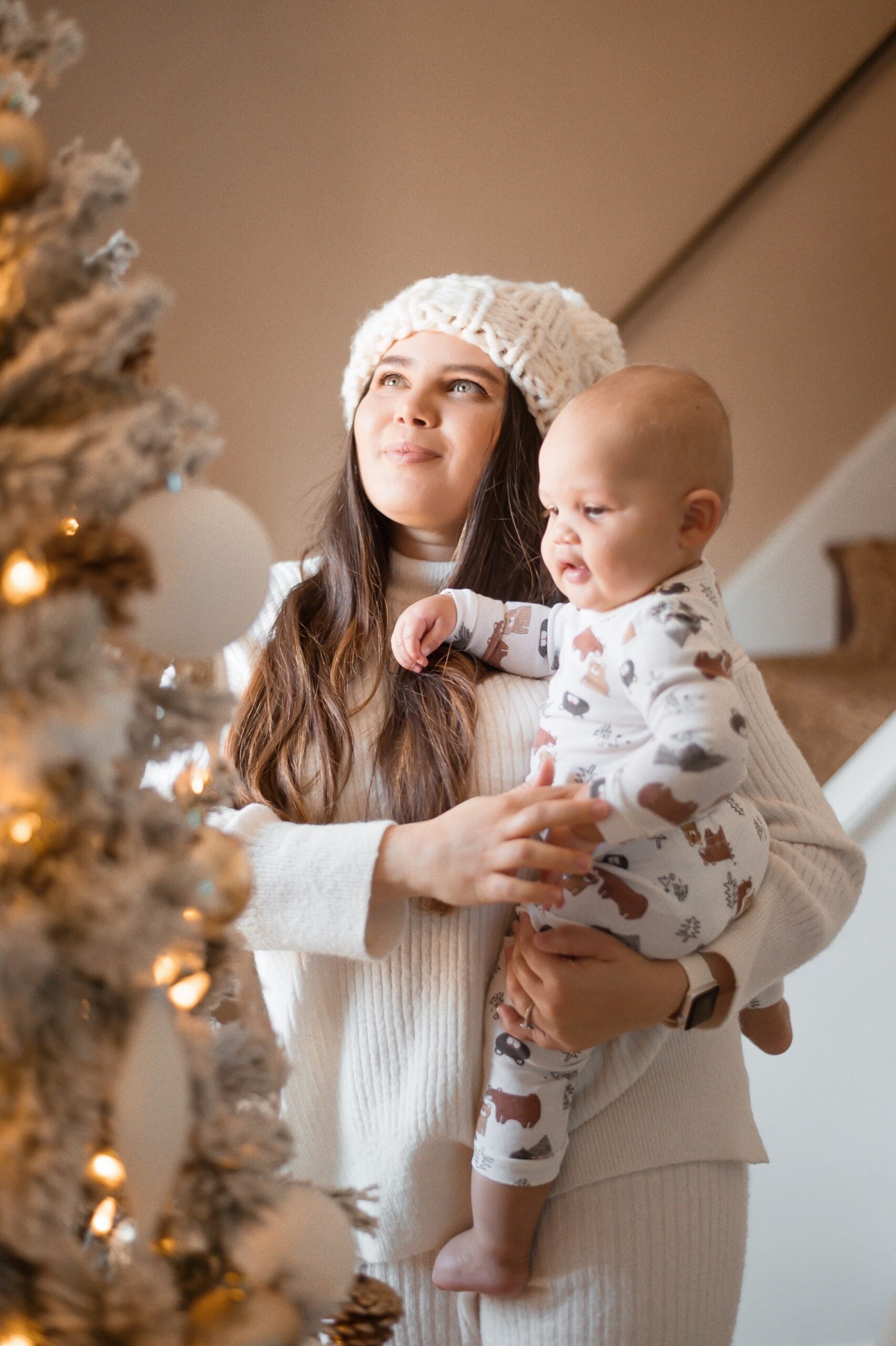 Mom wearing a cozy white sweater and knit hat holding her baby while they look at a decorated Christmas tree with soft warm lights.