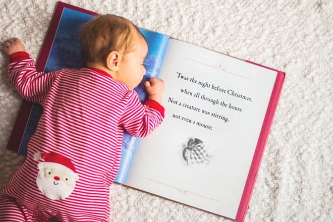 Baby in red striped Christmas pajamas lying on their tummy on an open book of “Twas the Night Before Christmas.”