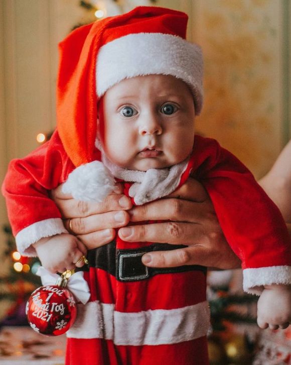 Baby dressed in a red Santa outfit being gently held upright, wearing a floppy Santa hat and holding a small red Christmas ornament.