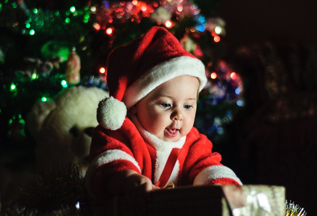 Baby dressed in a red Santa outfit, reaching toward a wrapped present with Christmas tree lights glowing softly in the background.