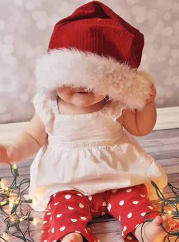 Baby sitting on the floor wearing a big Santa hat that covers their eyes, dressed in a white top and red polka-dot pants while holding a strand of Christmas lights.