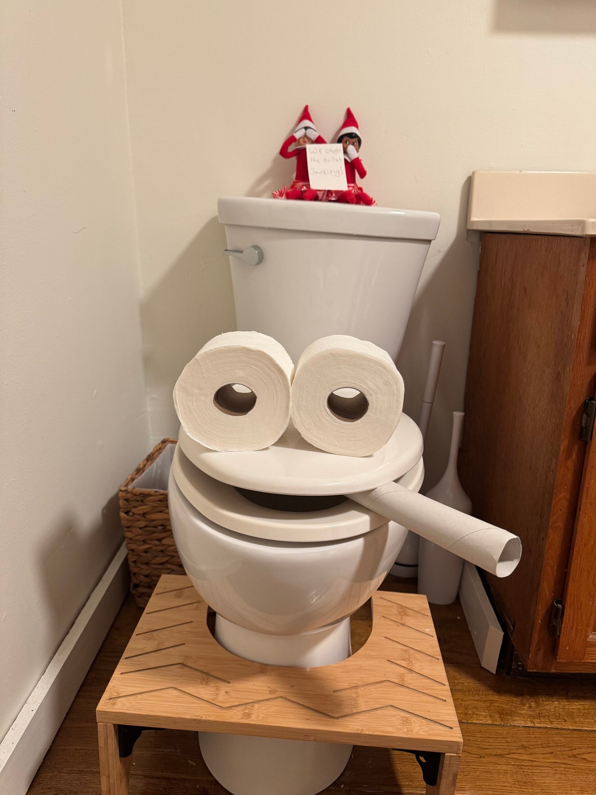 Funny Elf on the Shelf toilet smoking prank with toilet paper roll eyes and a cardboard tube cigarette while two elves sit on the tank.