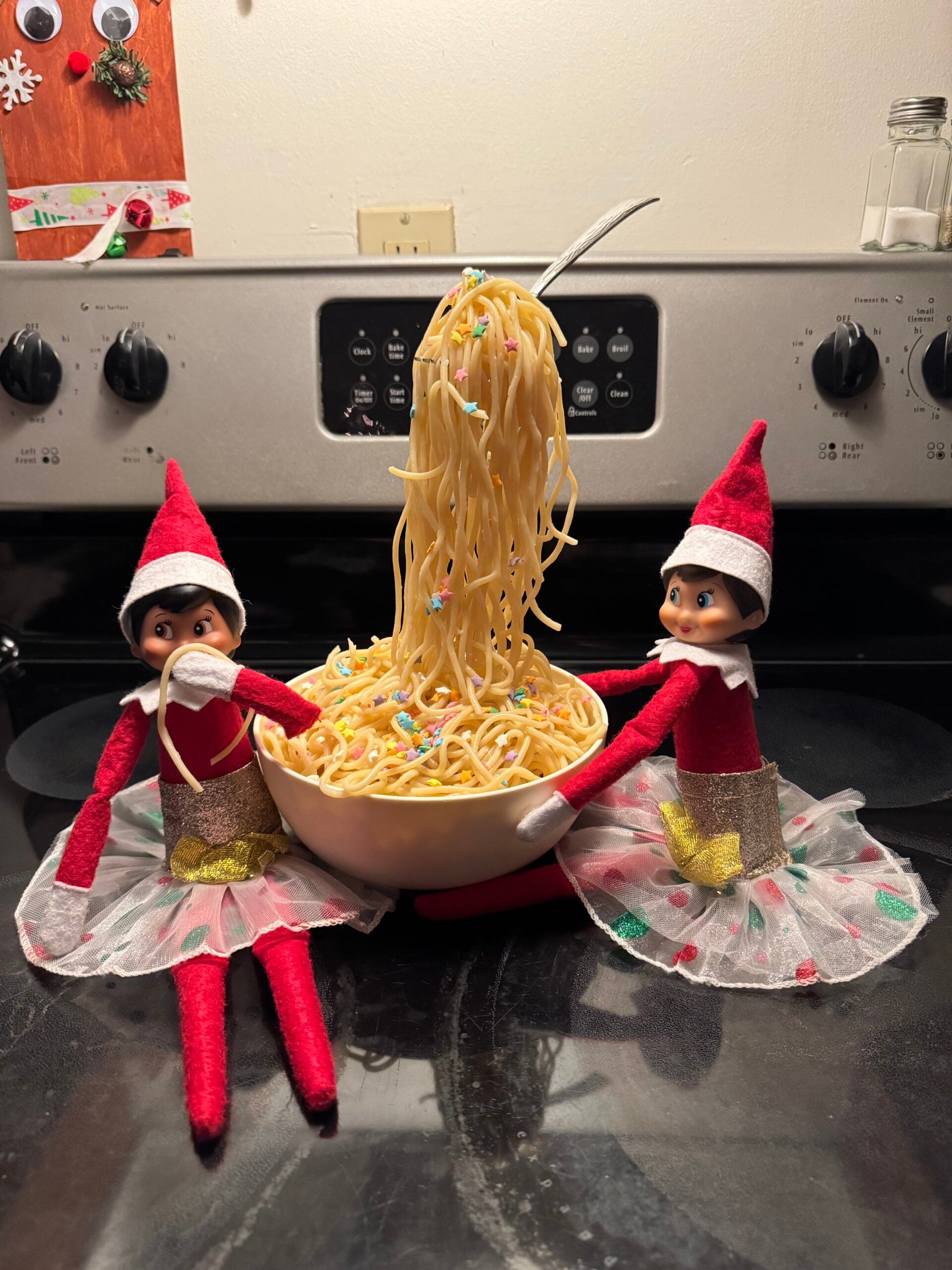 Funny Elf on the Shelf spaghetti trick with two elves beside a bowl of sprinkled spaghetti and a fork appearing to float above the bowl.