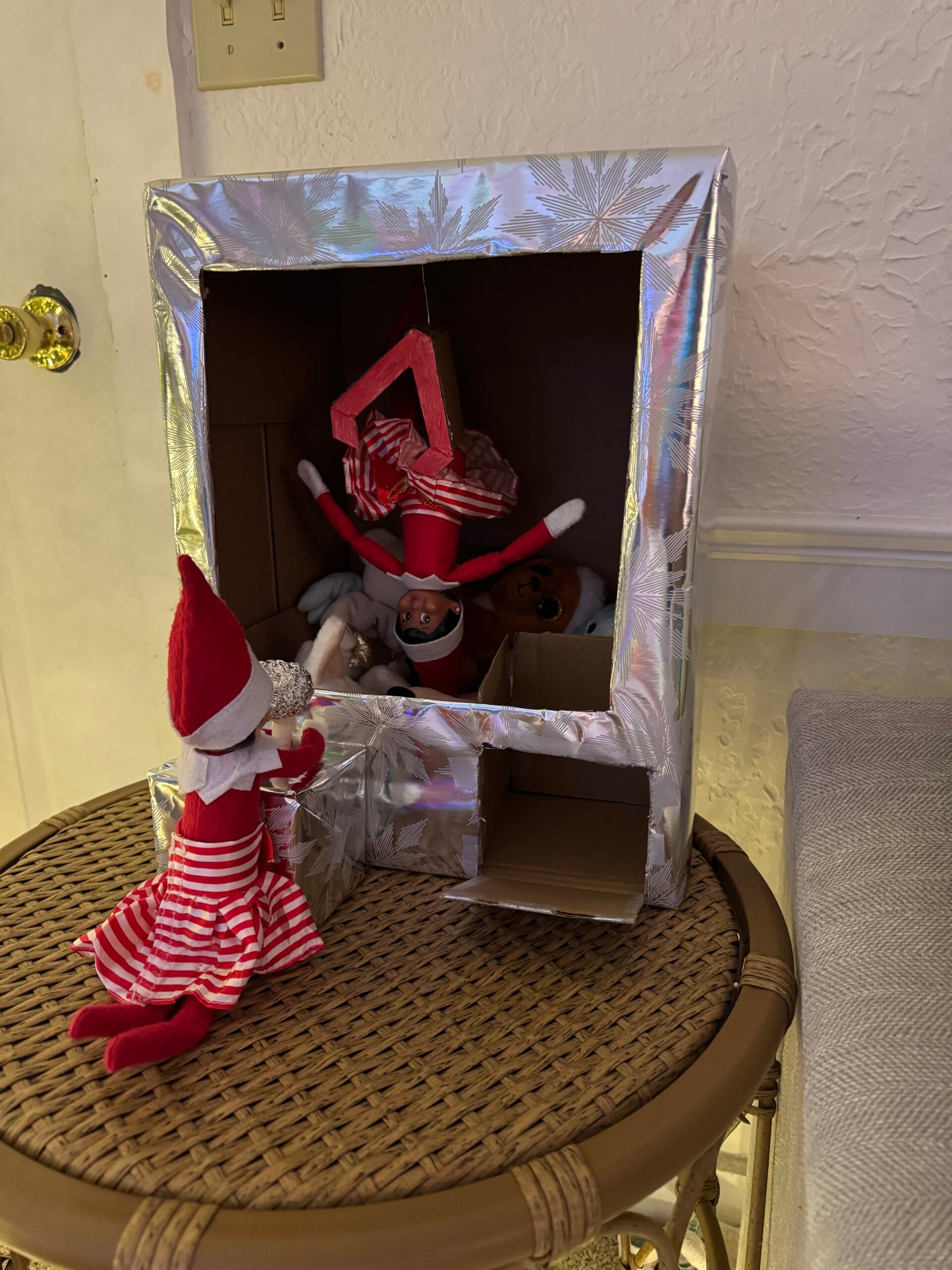 Creative Elf on the Shelf claw machine idea made from a cardboard box with one elf inside among stuffed animals and another operating the machine.