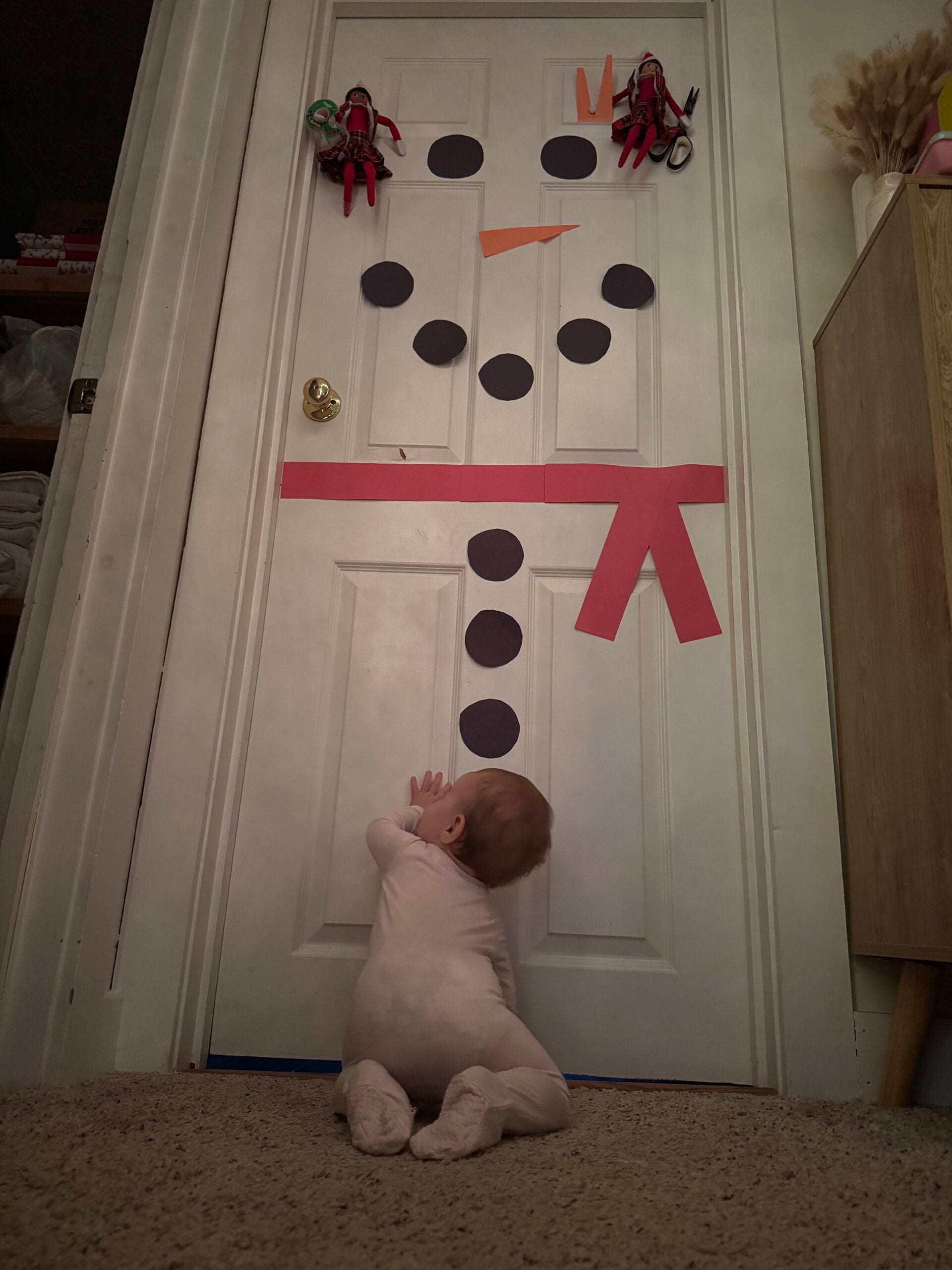 Easy Elf on the Shelf snowman door idea with two elves decorating a bedroom door with a carrot nose, coal buttons and red scarf.