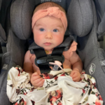Baby girl with pink headband buckled into a car seat.
