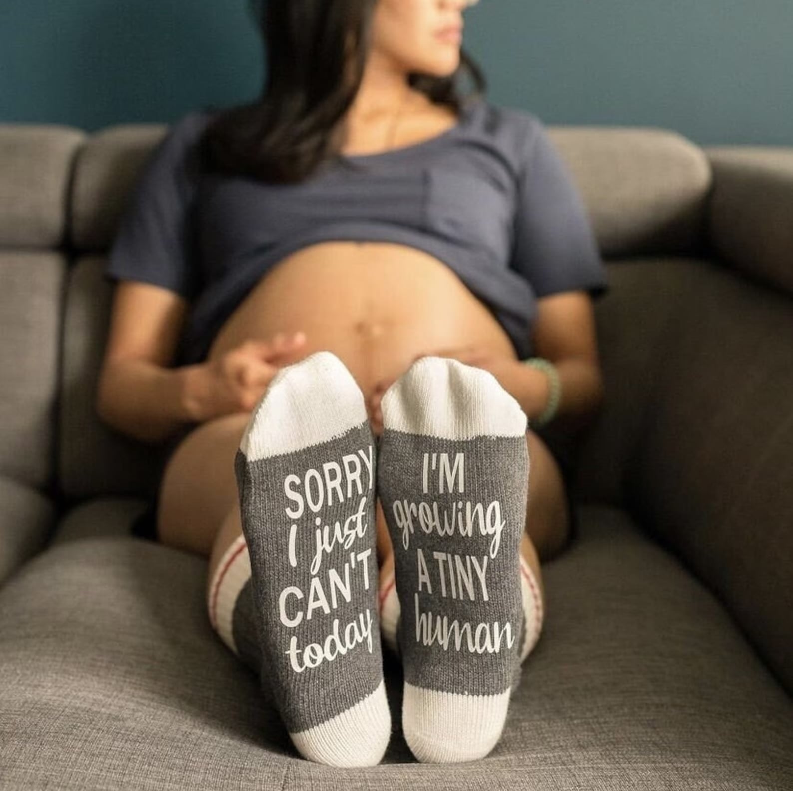 Pregnancy announcement photo idea featuring a pregnant mom sitting on a couch with her baby bump showing and gray socks that read “Sorry I just can’t today, I’m growing a tiny human.”