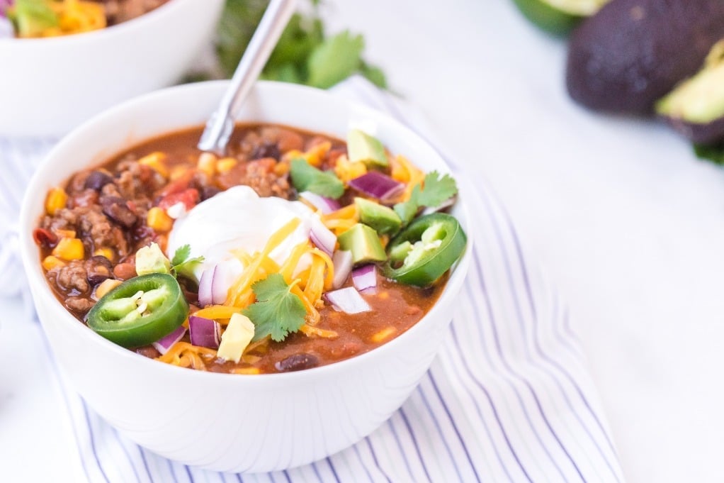 Bowl of crockpot taco soup topped with sour cream, shredded cheese, avocado, jalapeños, red onion, and cilantro for a crock-pot freezer meal for new moms.