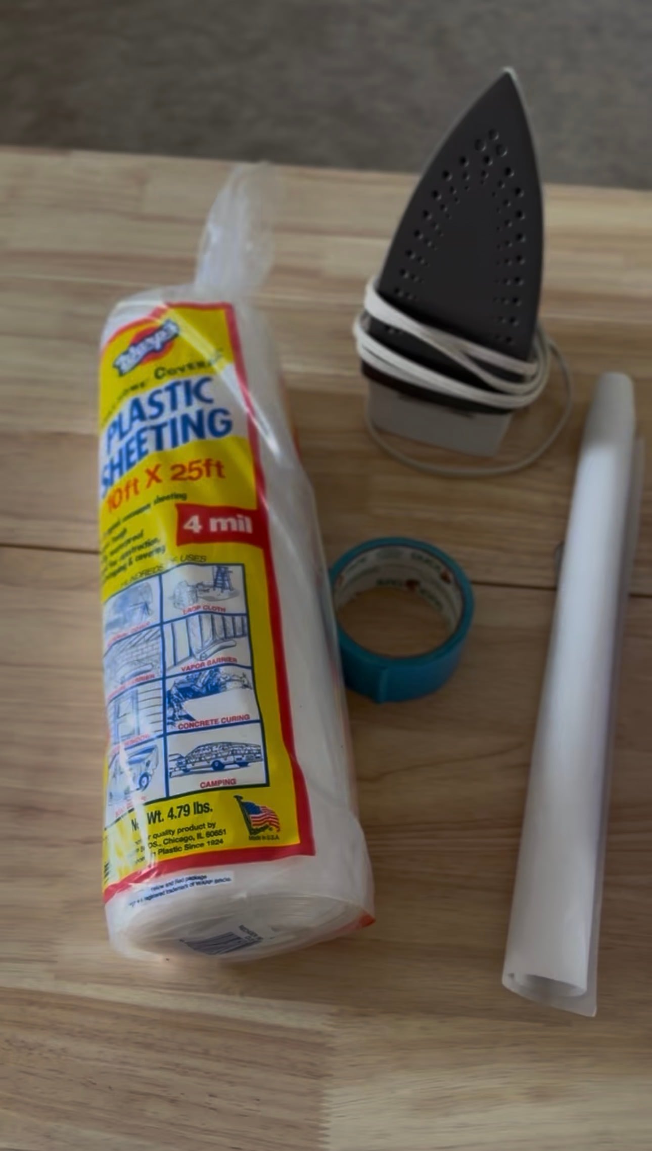 4 mil plastic sheeting, duct tape, parchment paper, and an iron laid out on a wooden table. Displayed as supplies needed to make a DIY water blob.