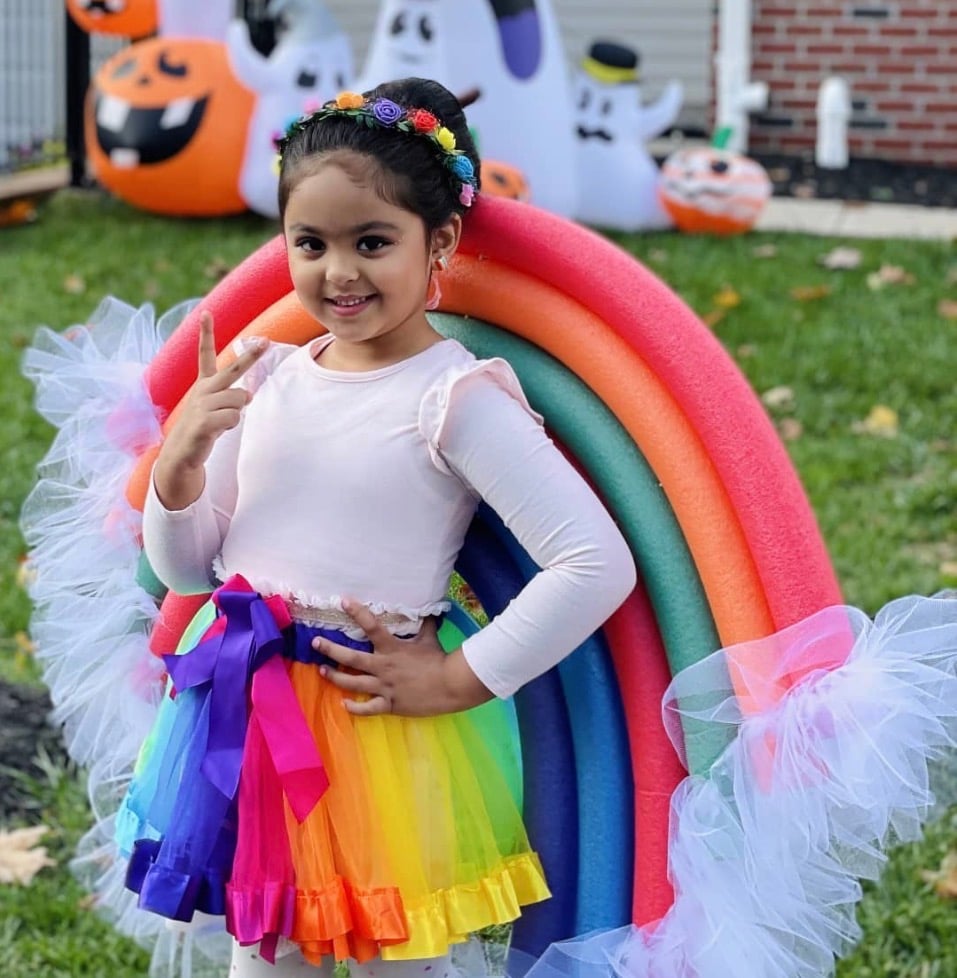 Rainbow DIY Halloween costume for toddler girl featuring colorful tulle skirt, rainbow pool noodle backdrop, flower headband, and white long sleeve shirt.