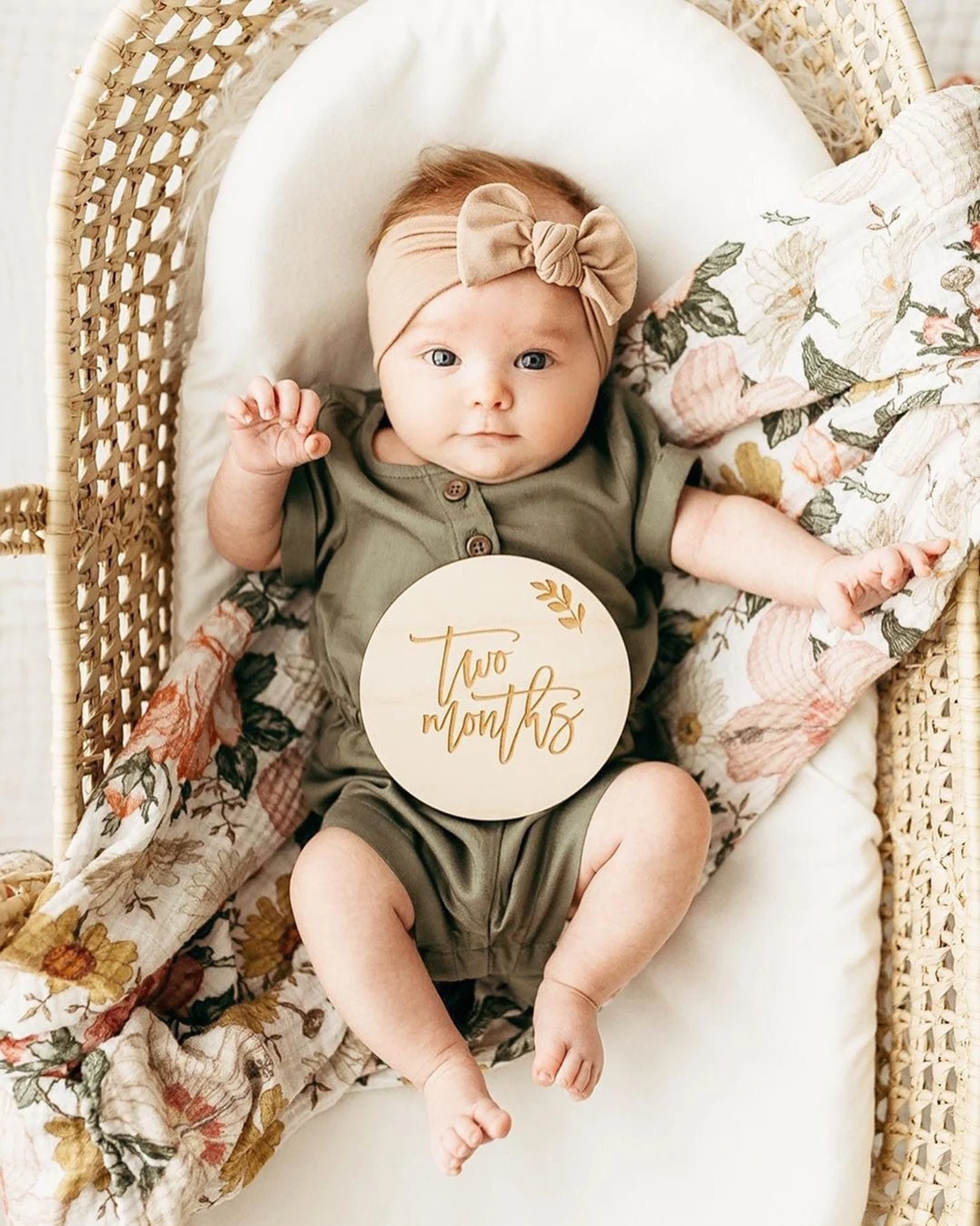 Baby in bassinet with floral blanket with wooden milestone marker in front of her.