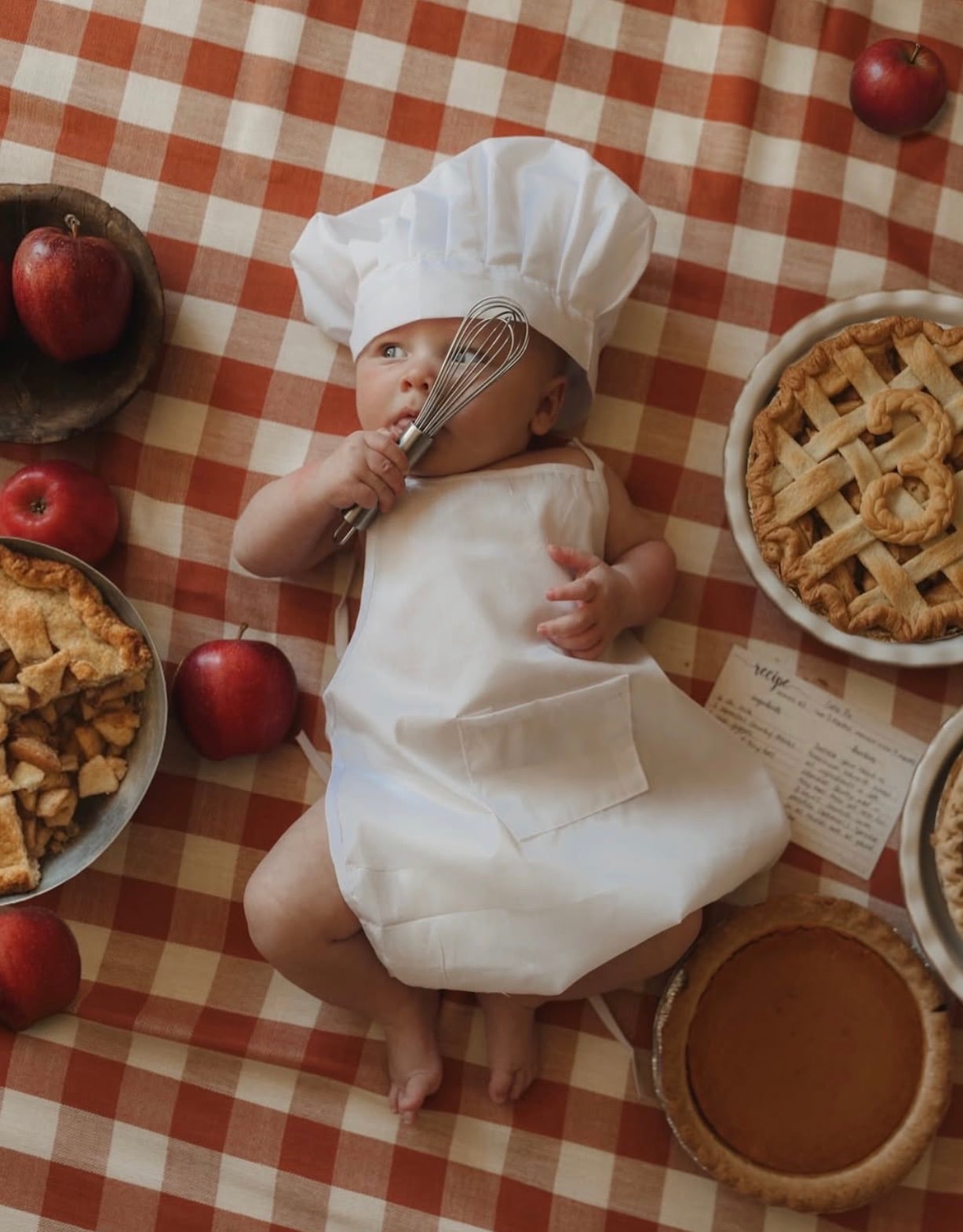 Monthly milestone photo of baby on red checkered blankets dress as a baker next to a pie with the number three on it.