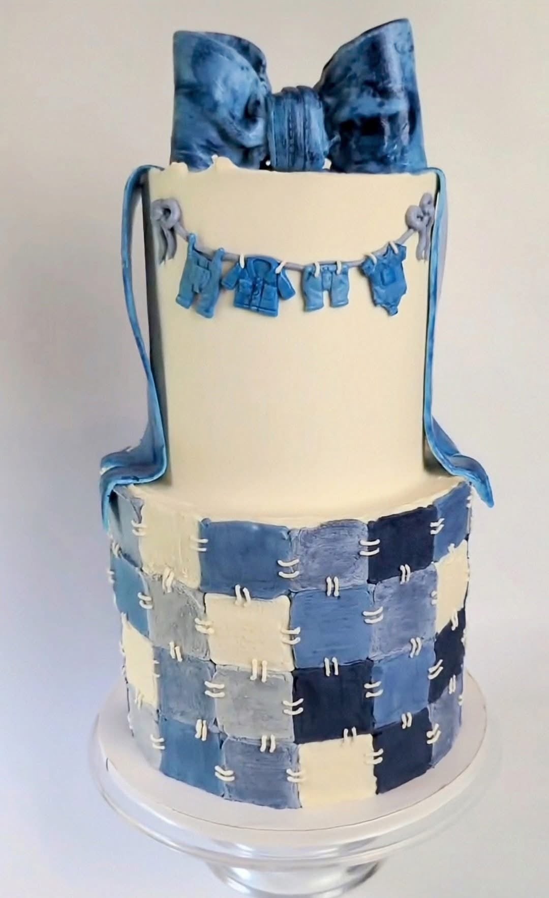 Two-tier Blue Jean Baby shower cake decorated with patchwork denim squares, tiny blue jean baby clothes, and a large blue bow for a blue jean baby shower theme.