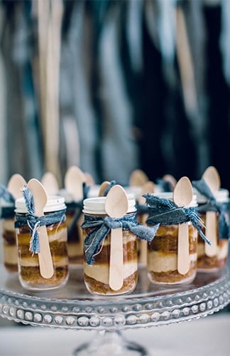 Individual layered desserts in small mason jars with wooden spoons and denim fabric bows for a blue jean baby shower theme.